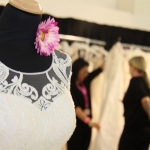 Pressebereich der Hochzeitsmesse HOCHZEITSTAGE, weißes Hochzeitskleid mit durchsichtiger Spitze am Ausschnitt, pinke Blume am Hals der Schneiderpuppe, Brautkleidberatung im Hintergrung
