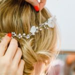 Hochzeitstage, Hände mit rotem Nagellack setzen einer jungen Frau filigranen Schmuck ins Haar mit weißen Blüten und Perlen