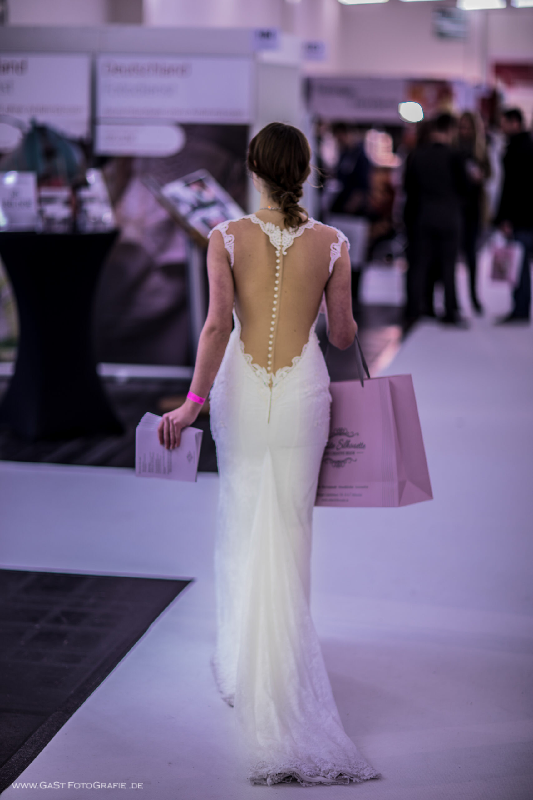eine junge Frau in weißem Brautkleid mit tiefem Rücken läuft die Gänge einer Messehalle entlang und verteilt Flyer